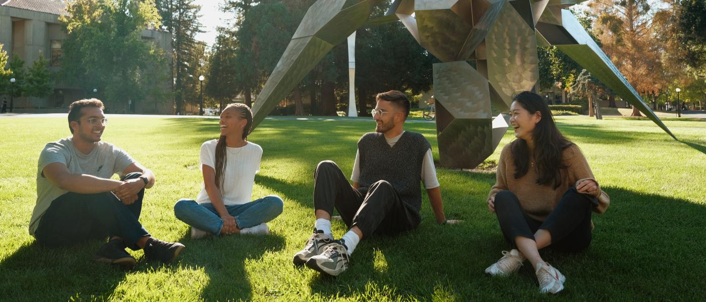 Four students talking and sitting on a grassfield in front of a large artwork.