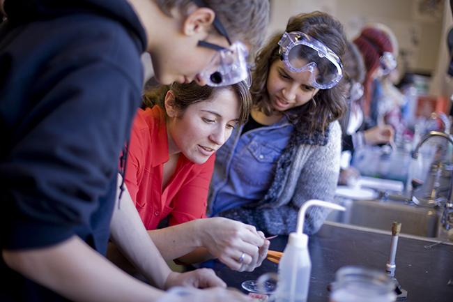 Teacher demonstrating a lab task to two students wearing lab goggles.