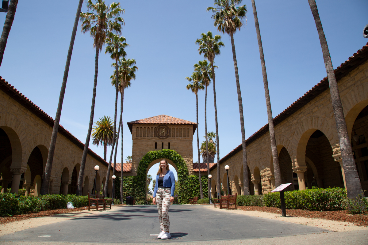 Photo of Gerta Guitart, standing, with palm trees and the Stanford Main Quad behind her