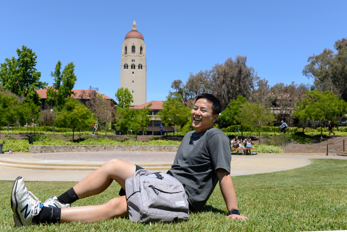 Photo of Richard Tang sitting on the grass in the sunshine, with Stanford's Hoover Tower in the background