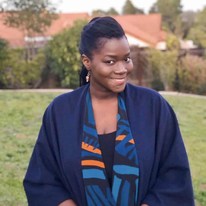 Picture of Diarra Bousso, outside, smiling, wearing a blue draped cape and a vibrant, colorful scarf with a bold, graphic print in black, blue, and orange