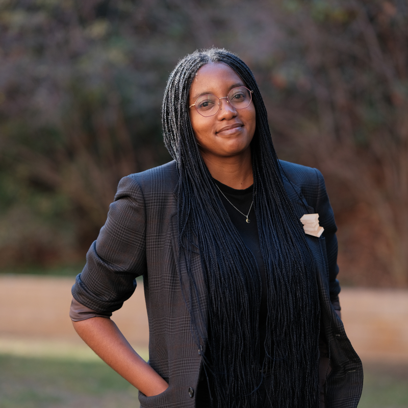 Photo of Geraldine Mukumbi standing outside, smiling, with a pin of a stack of books on her lapel.