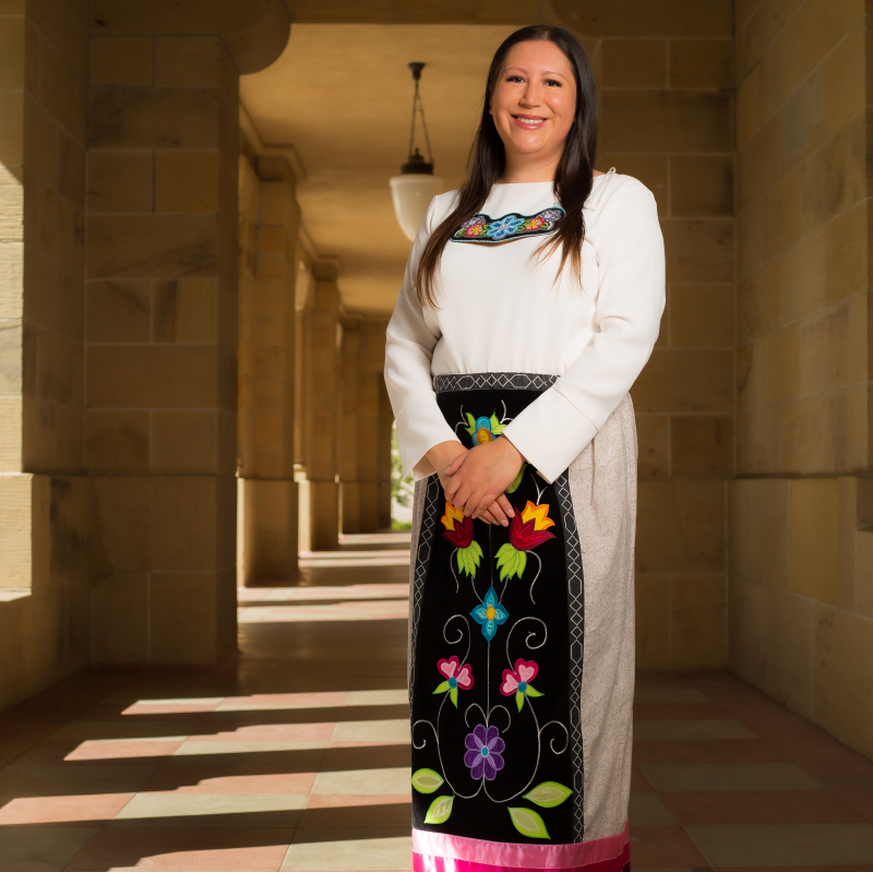 Jarita Greyeyes, standing in the arcade of a Stanford sandstone building, wearing a ribbon skirt with Anishnaabe florals
