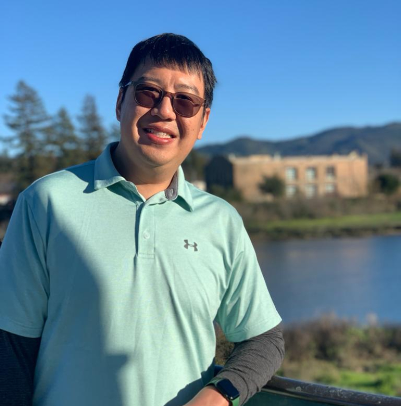 Chee Wing Chan standing outside, smiling, with a lake, a building and some hills in the background