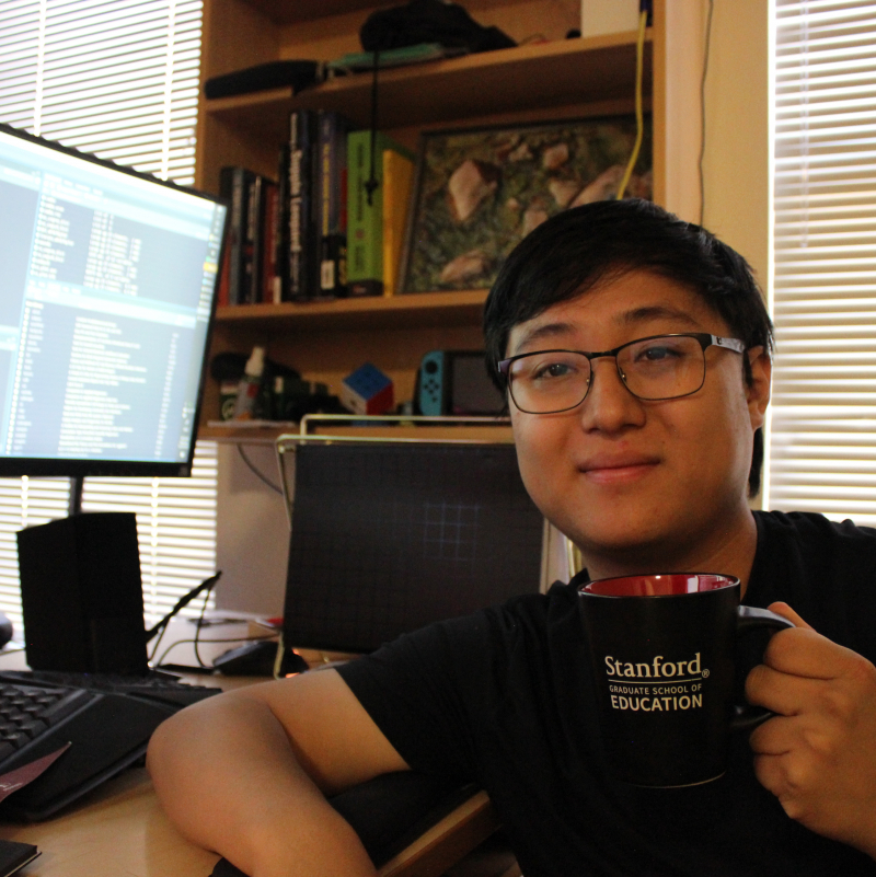 Raymond Zhang at his desk, holding a Stanford GSE mug in front of a monitor with coding