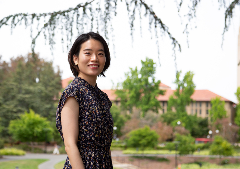 Photo of Risa Ninomiya standing outside on the Stanford campus, with leafy trees and buildings with red tiled roofs in the background
