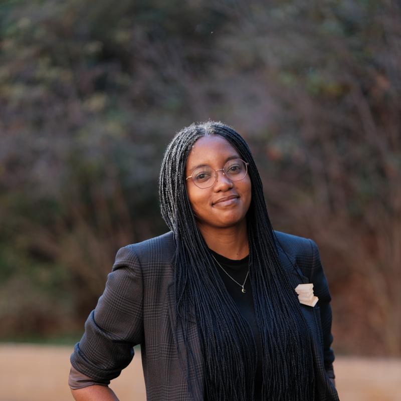 Photo of Geraldine Mukumbi standing outside, smiling, with a pin of a stack of books on her lapel.