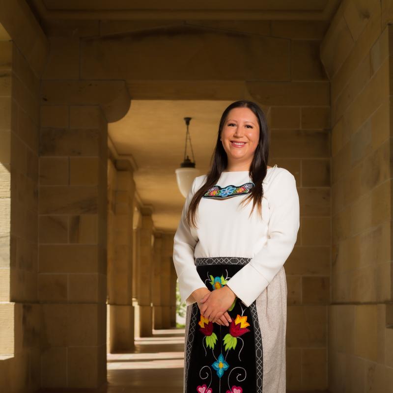 Jarita Greyeyes, standing in the arcade of a Stanford sandstone building, wearing a ribbon skirt with Anishnaabe florals