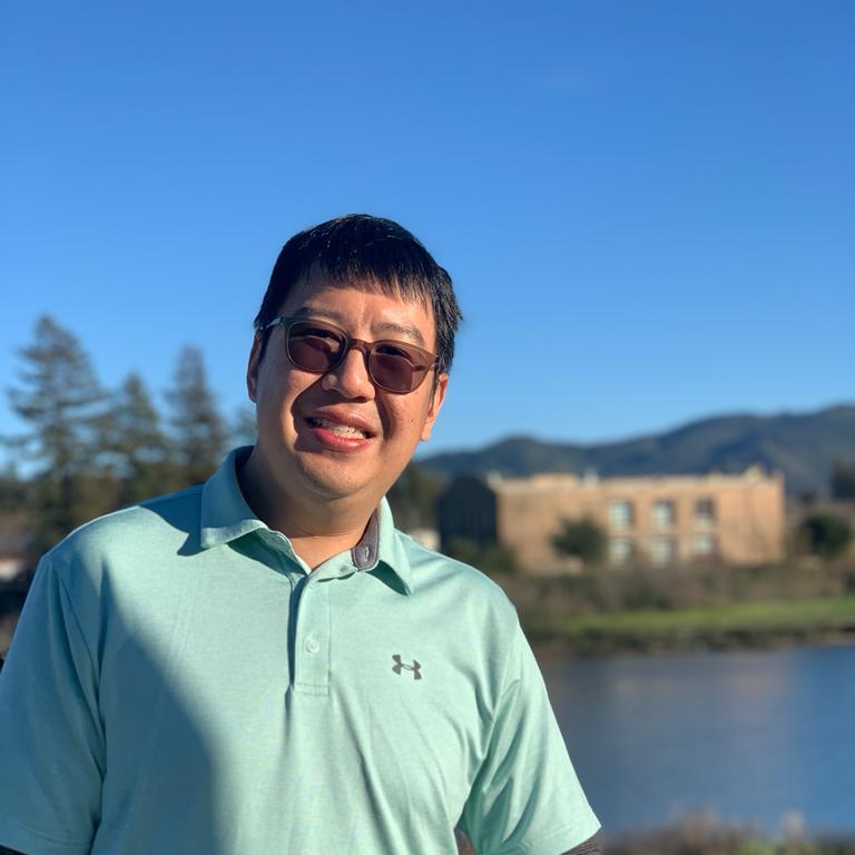 Chee Wing Chan standing outside, smiling, with a lake, a building and some hills in the background