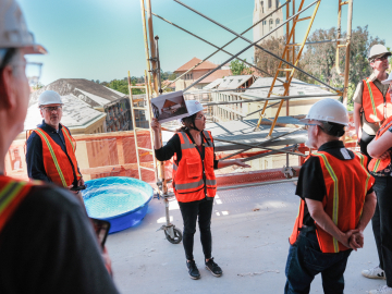 Photo of a group of people, all wearing bright orange construction vests and hard hats, on the top floor of a tall construction site with a view of the old education building and Hoover Tower in the background